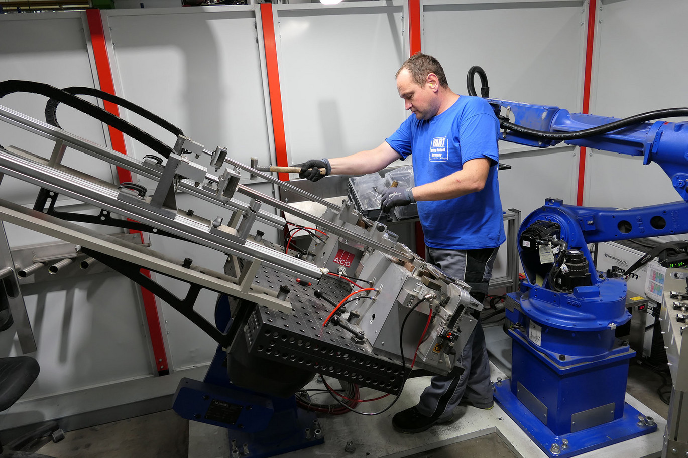 Person in blauer Uniform bedient eine industrielle Montageanlage mit Roboterarm in einer Werkstatt, während sie eine Komponente justiert