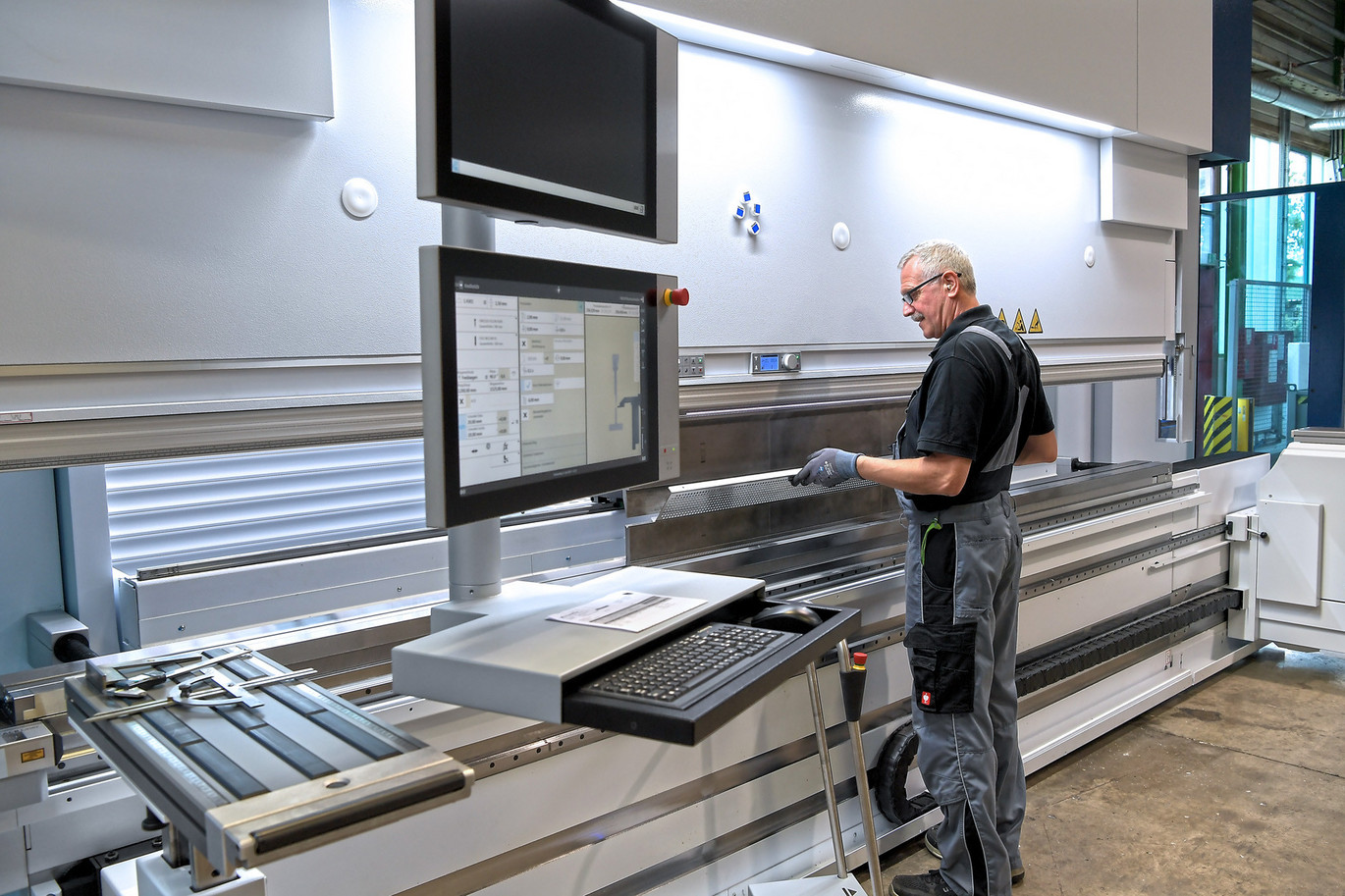 Person bedient eine moderne CNC-Biegemaschine in einer sauberen, gut beleuchteten Werkstatt, um ein Metallstück zu bearbeiten, mit zwei Steuerungsmonitoren